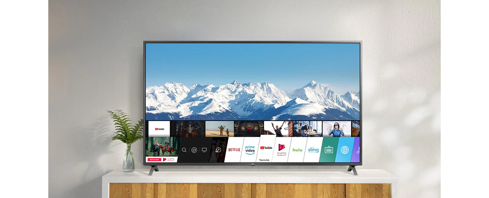 A TV em um suporte branco contra uma parede branca. A tela da TV mostra a tela inicial com o sistema operacional webOS.